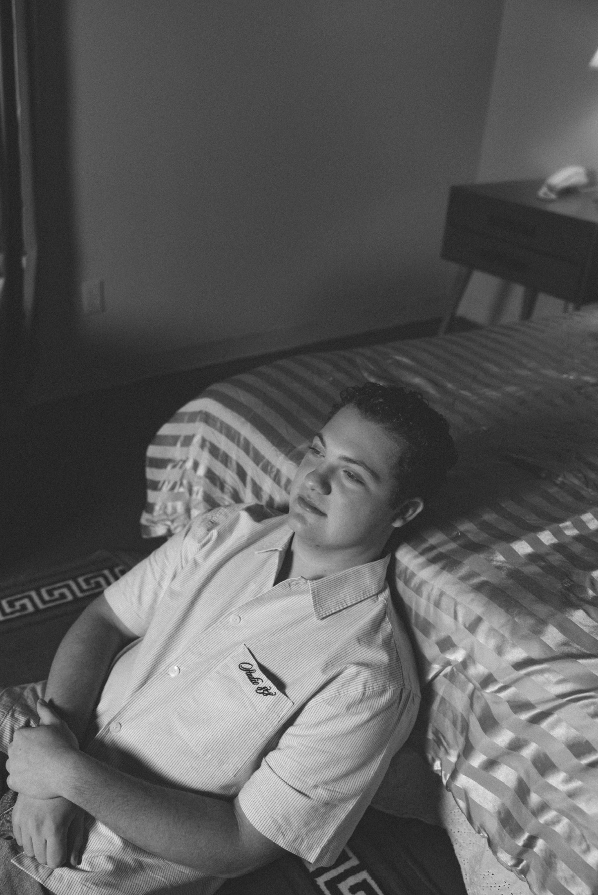 Black and white photo of Davey J leaning against a bed in a striped room